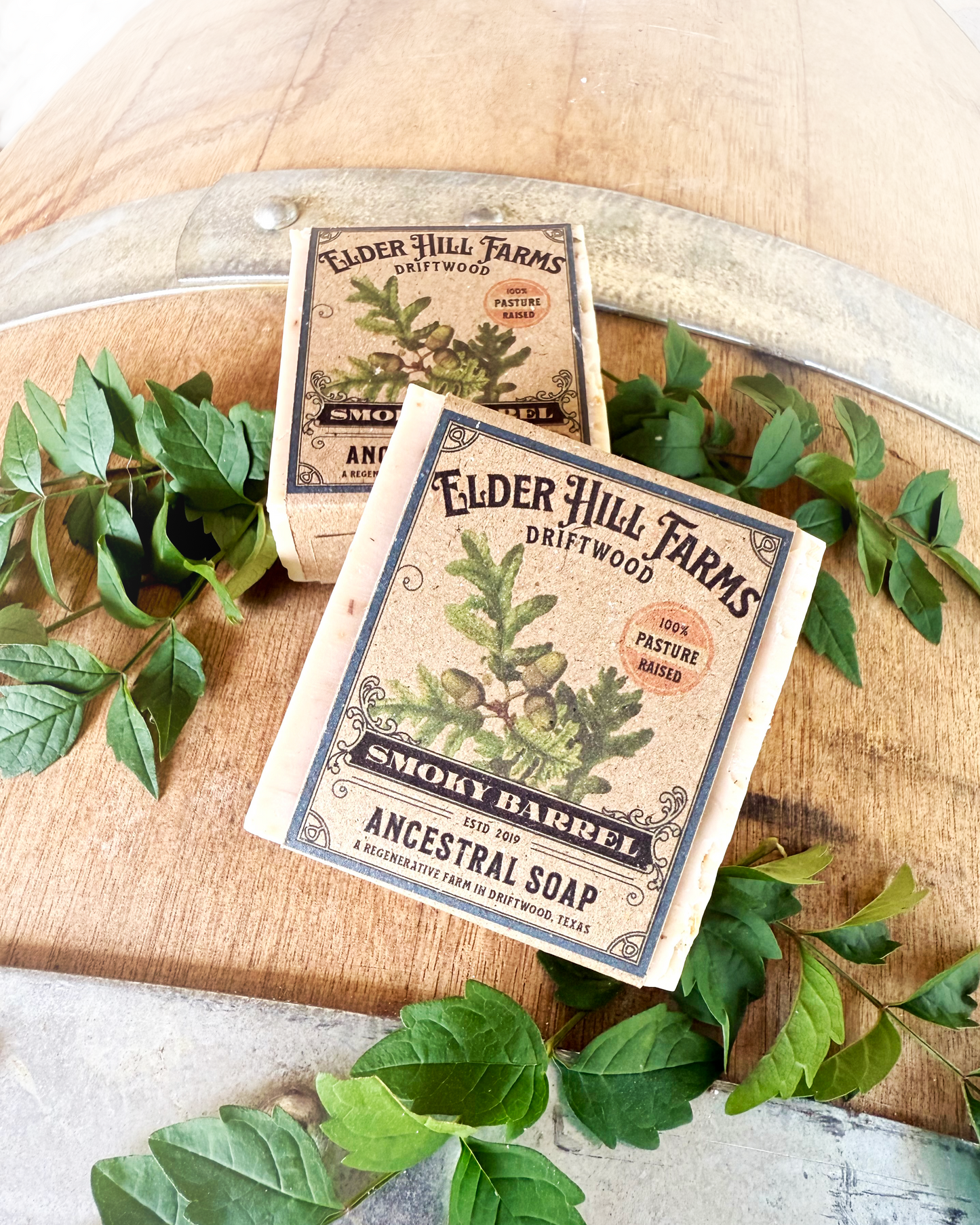 Two packages of Elder Hill Farms soap on a wooden barrel surface with green leaves.