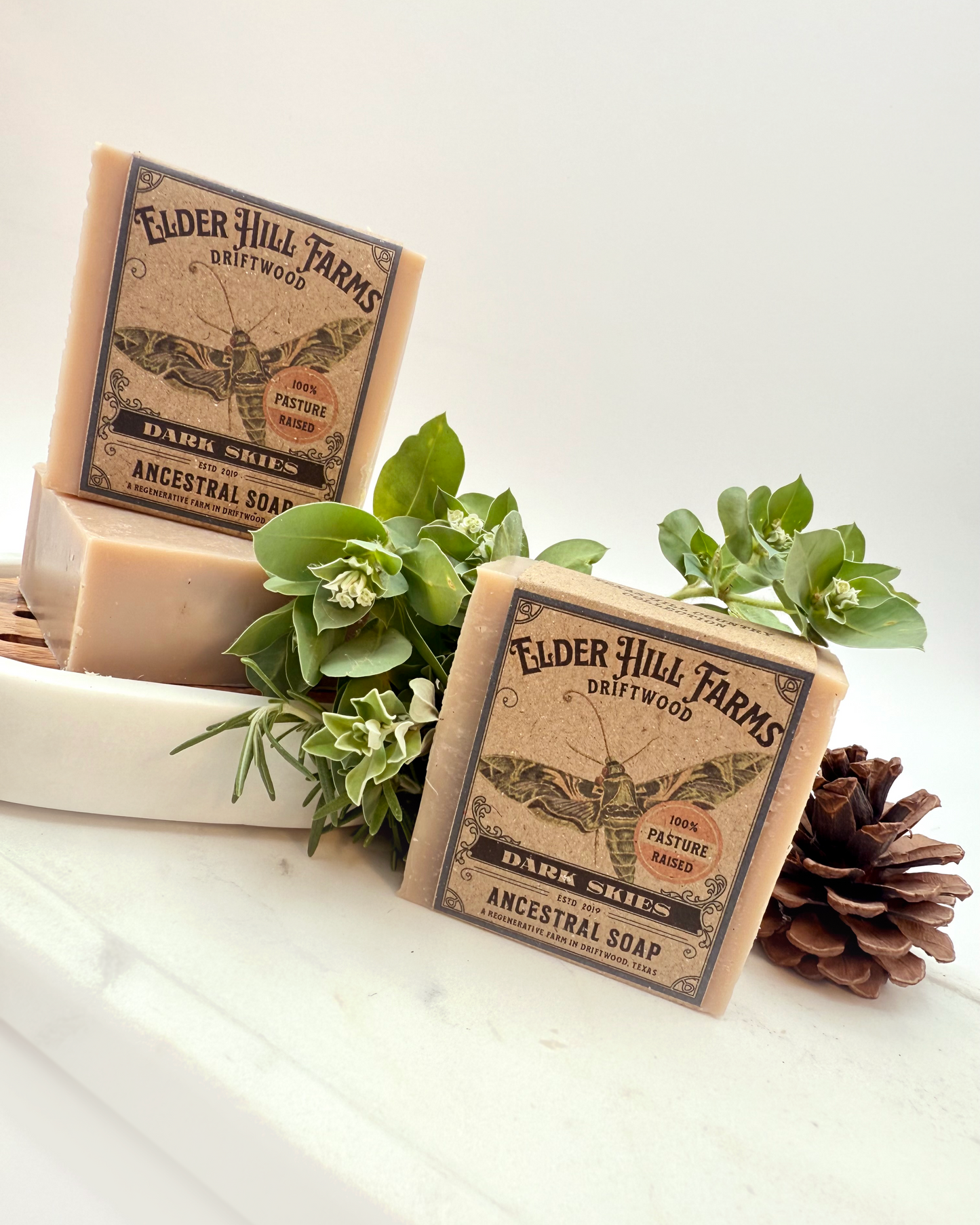 Two bars of Elder Hill Farms soap with packaging on a white surface with greenery.