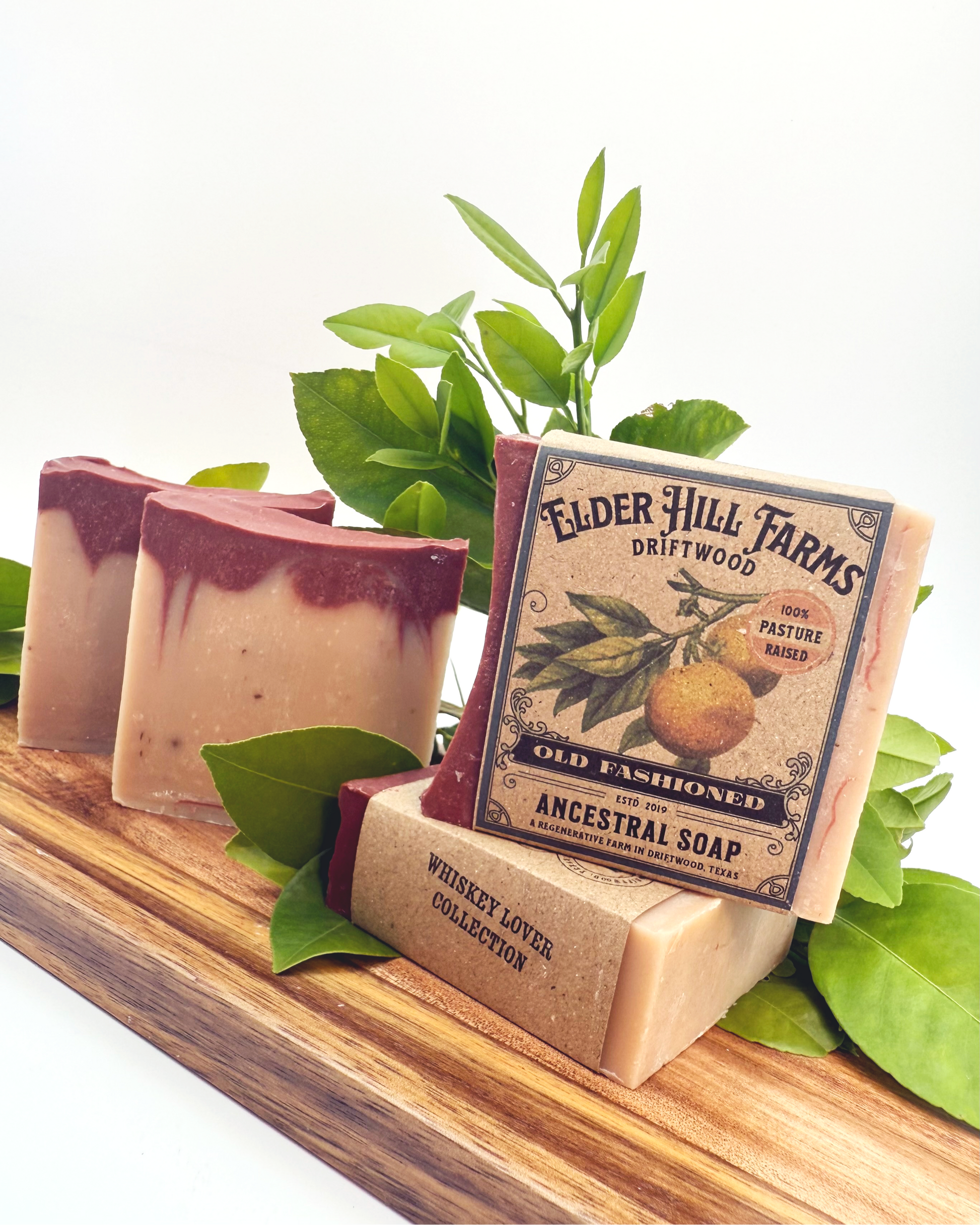 Three bars of soap on a wooden stand with a 'Elder Hill Farms' label, surrounded by green leaves.